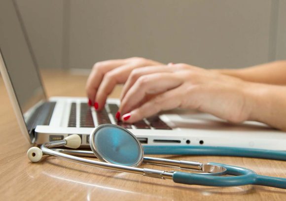 A case manager types on a laptop with a stethoscope on the table. Close up.