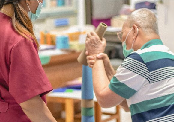 An occupational therapist works with an older patient on grip strength and arm mobility. Both are masked.