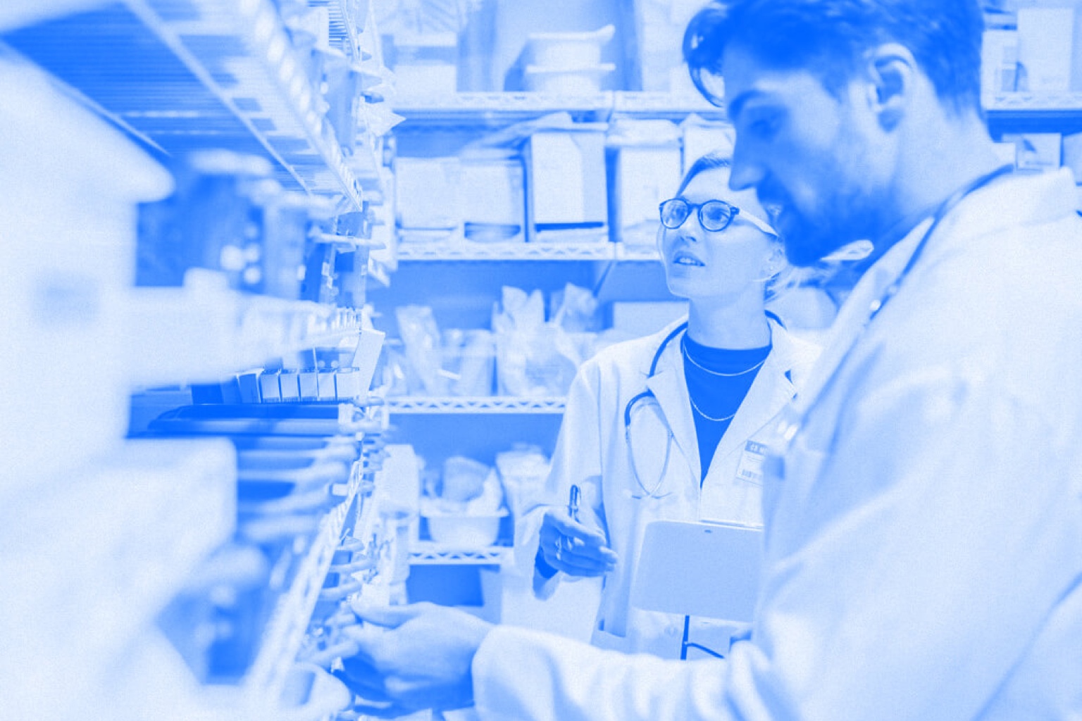 A pharmacy technician and pharmacist work together while looking at shelves of medicine. Blue-tinted photograph.