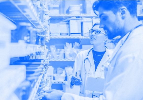 A pharmacy technician and pharmacist work together while looking at shelves of medicine. Blue-tinted photograph.