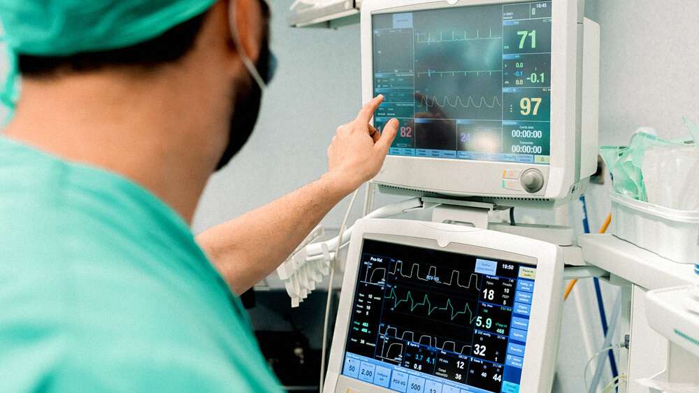 A nurse monitors a patient’s vitals from two monitors—pointing at one specific graph.