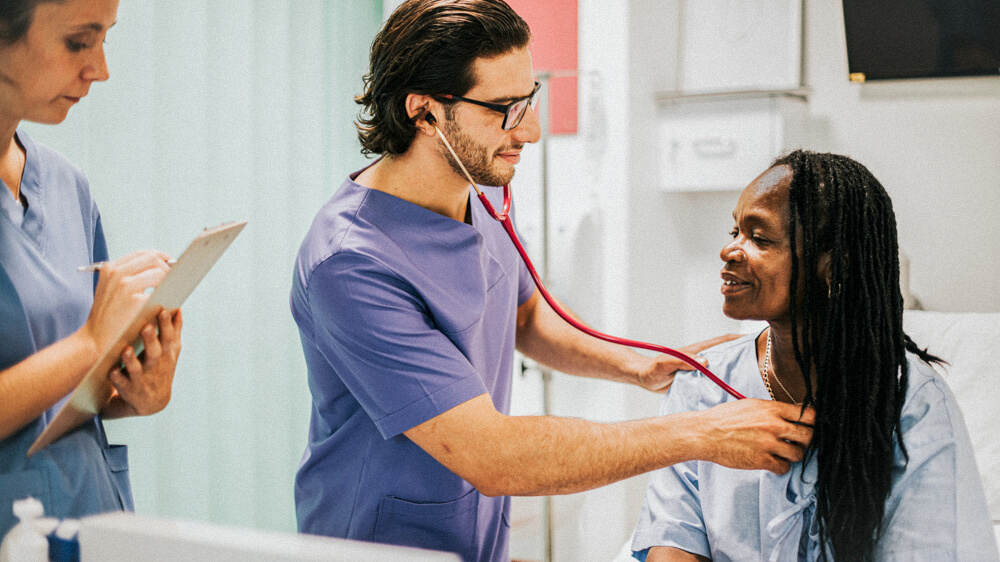 Two nurses in a hospital room with a patient. One nurse holds a stethoscope to the patient’s chest.