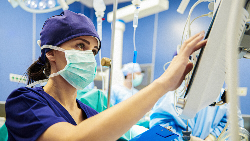 A masked nurse reviews a monitor while assisting in a surgery room.