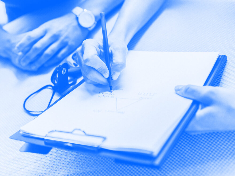 Hands writing a note on a notepad held out on a desk next to another person.