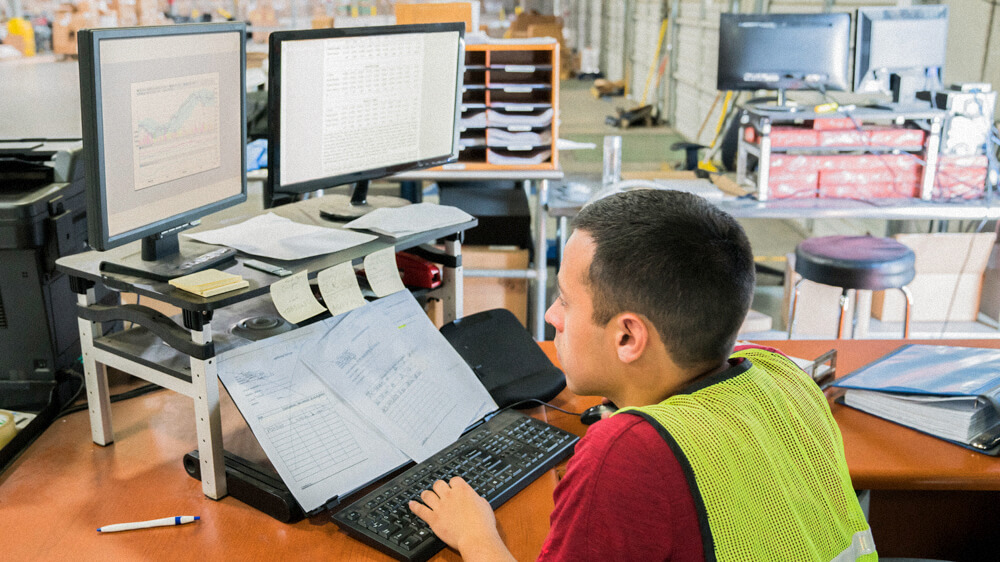 A person wearing a safety vest works at a desk with two monitors, sticky notes, and printouts.