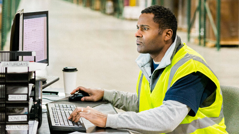 A person sits at a desk in a warehouse, looking at their computer.