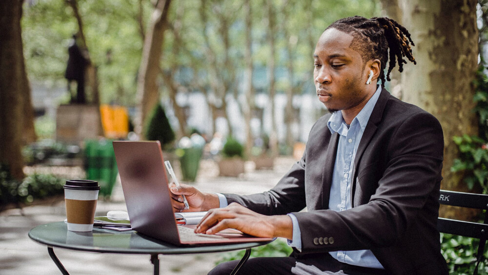 A person works on their laptop from a cafe table in the park.