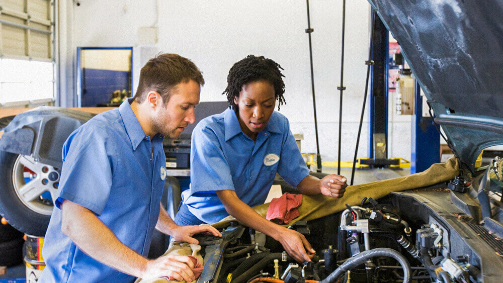 Two automotive mechanics work under the hood of a vehicle together.