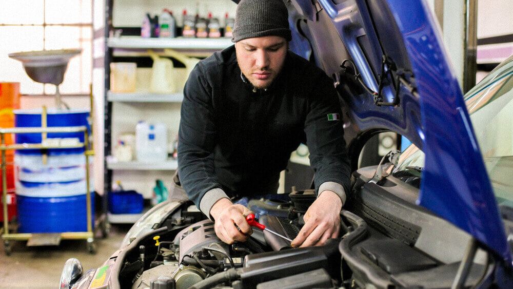 Automotive mechanic working under the hood of a blue vehicle in a shop.