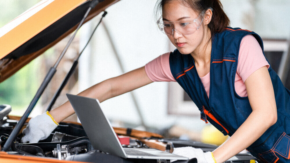 Automotive mechanic adjusting something under the hood while referencing a laptop.