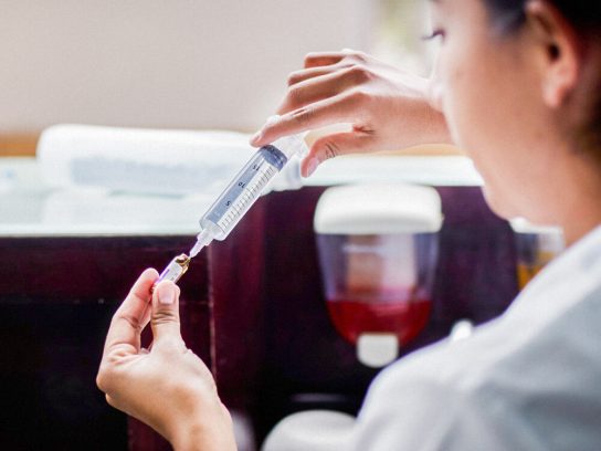 Hands measure medication from a vial with a patient and nurse in the background.