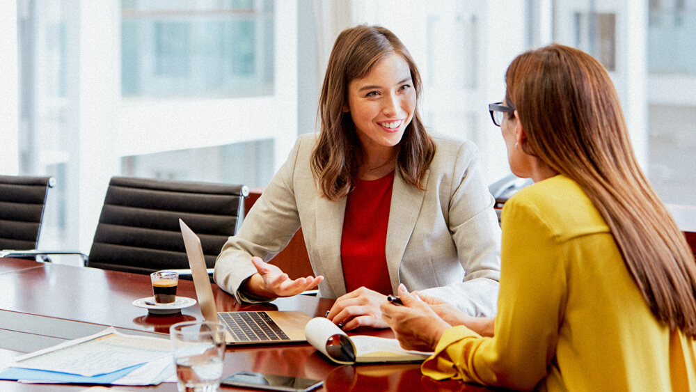 A certified financial planner talks with a client in a corporate office over laptop and notes.