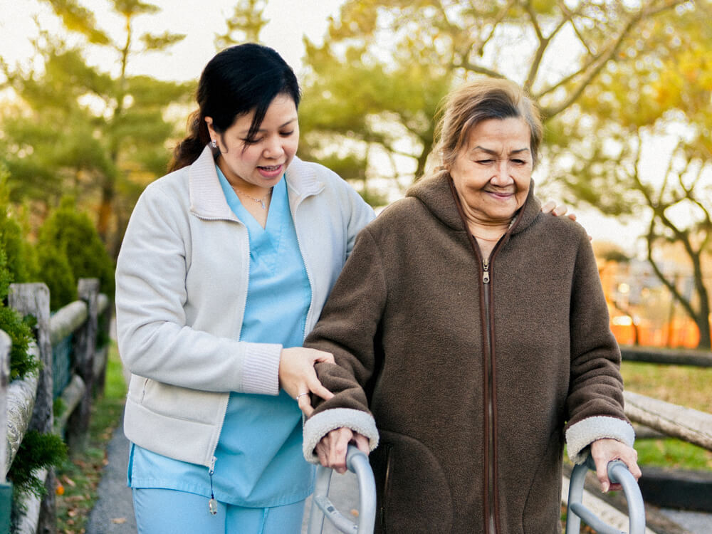 A nursing assistant helps a person walk outside with a walker.