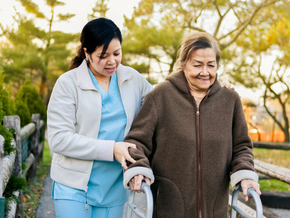 A nursing assistant helps a person walk outside with a walker.