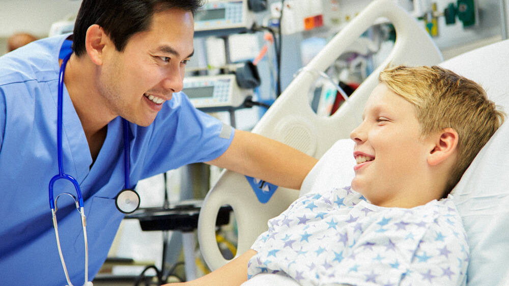 A nursing assistant leans in and talks with a child patient on a medical bed.