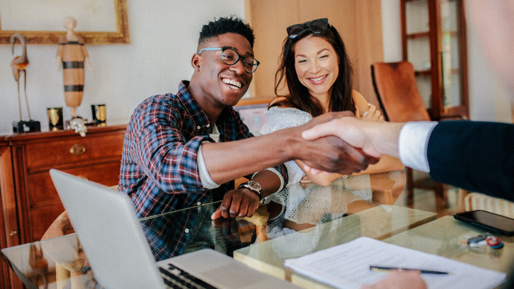 A realtor shakes hands with a couple from across their desk.