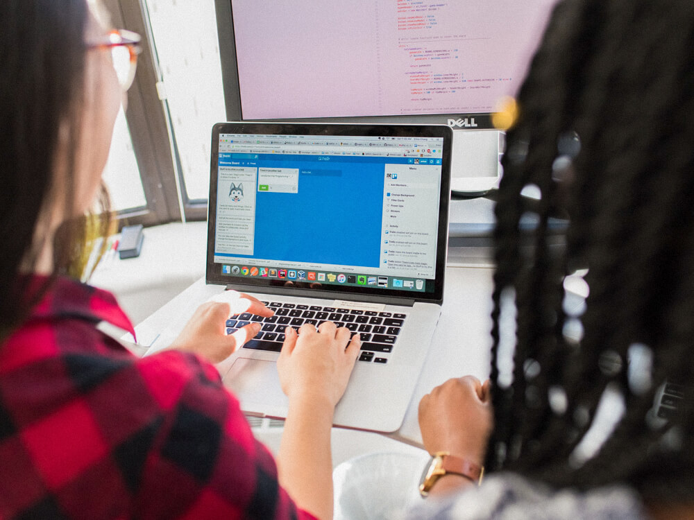 Two project managers work together looking at a laptop and a monitor screen.