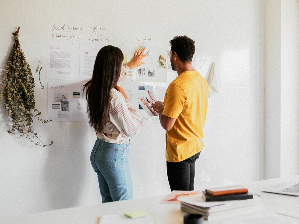 Two professionals review work posted on the wall.