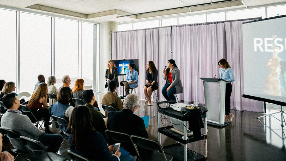 A panel of speakers sits in front of an audience in a conference setting.