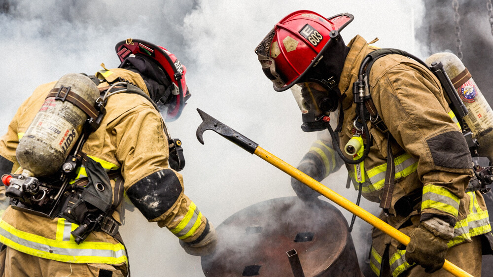 Two firefighters in full gear carry a metal drum in a scene of smoke.