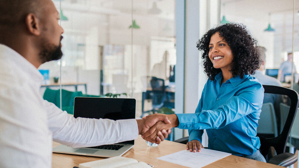 Two people shake hands over a desk in an office.