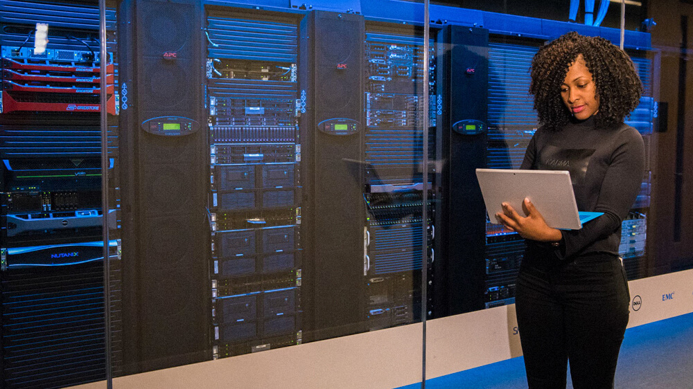 Information security manager stands in front of servers while monitoring from a laptop.