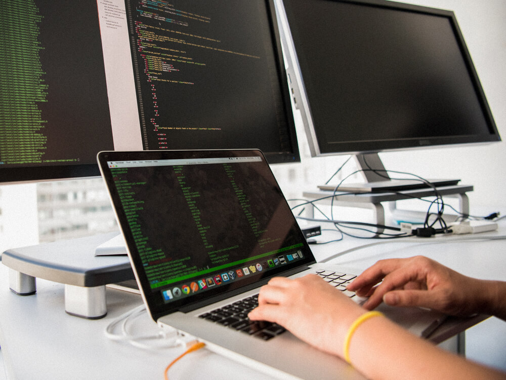 Hands typing on a laptop keyboard at a workstation with three monitors.