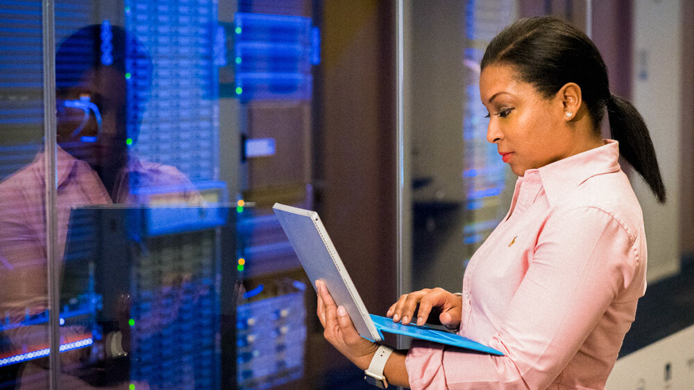 Cloud security professional stands in front of servers while monitoring from a tablet.