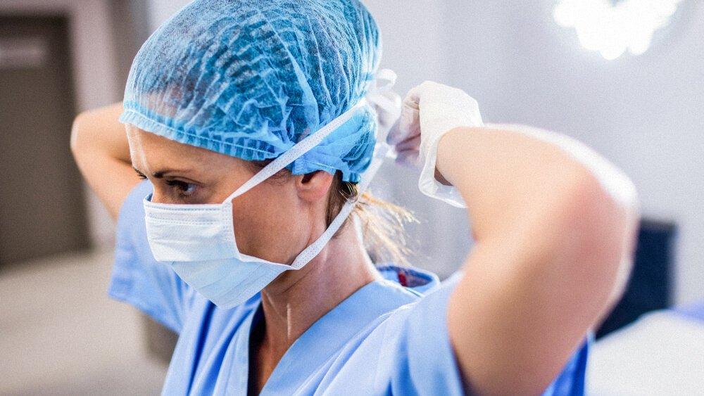 A nurse wearing a head covering and surgical gloves ties a mask over their face.