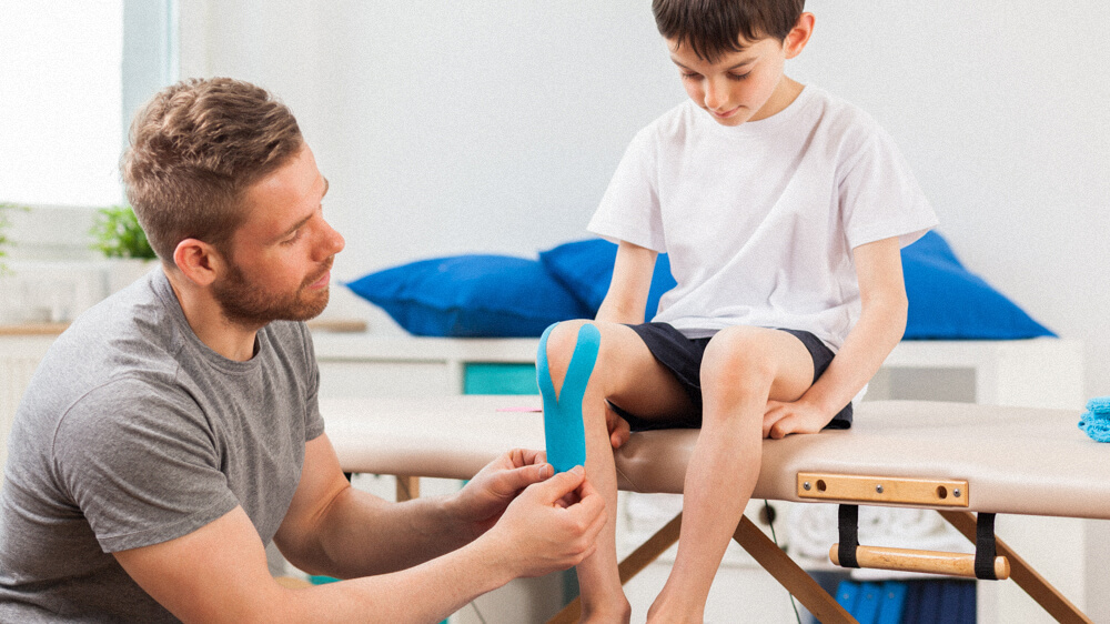 An occupational therapist assistant applies medical tape around a child’s knee.