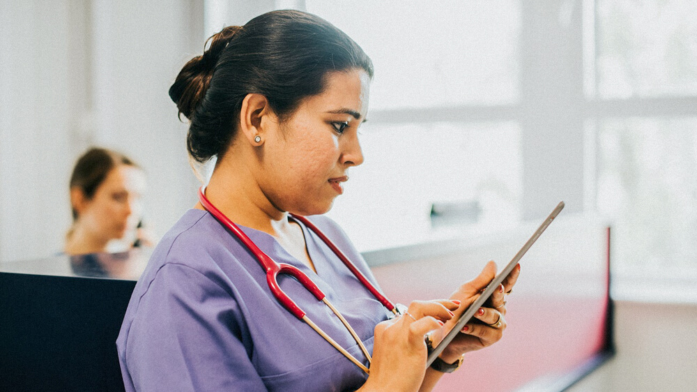 A nurse wearing a stethoscope reads from a tablet.