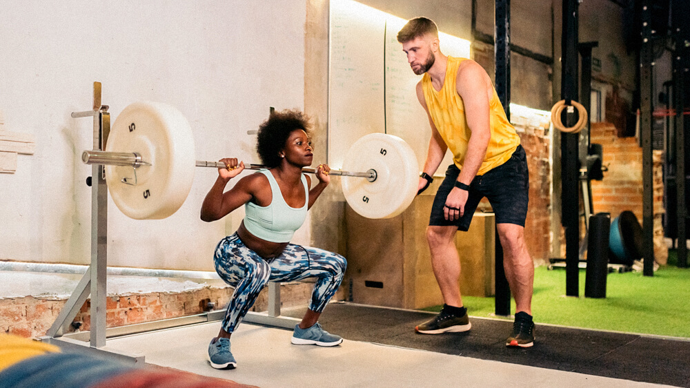 A personal trainer monitors a client performing weighted barbell squats.