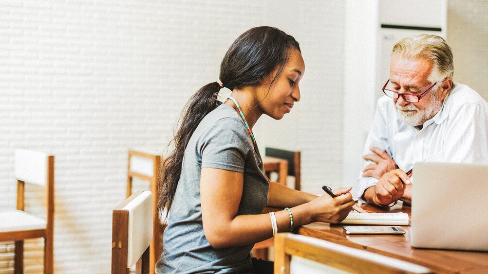 A student meets with their instructor at a desk in a classroom.