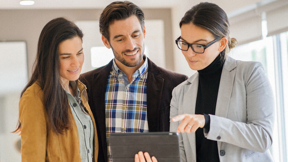 A realtor stands with a couple and holds a tablet for them to review together.