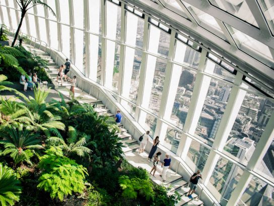 Inside of a building with a high glass ceiling, lots of green plants, and people walking along a walkway.