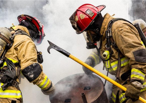 Two firefighters in full gear working to move a barrel while holding a hook.