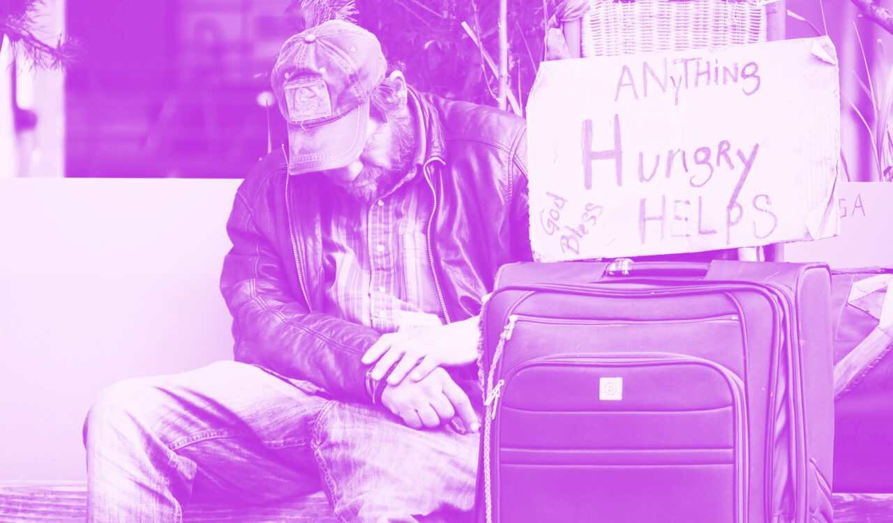 Homeless person sitting next a suitcase and sign asking for help.