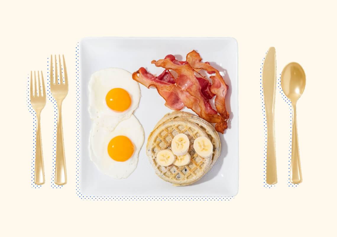 Bacon, eggs, waffles, and cut bananas on a square plate with silverware on either side.