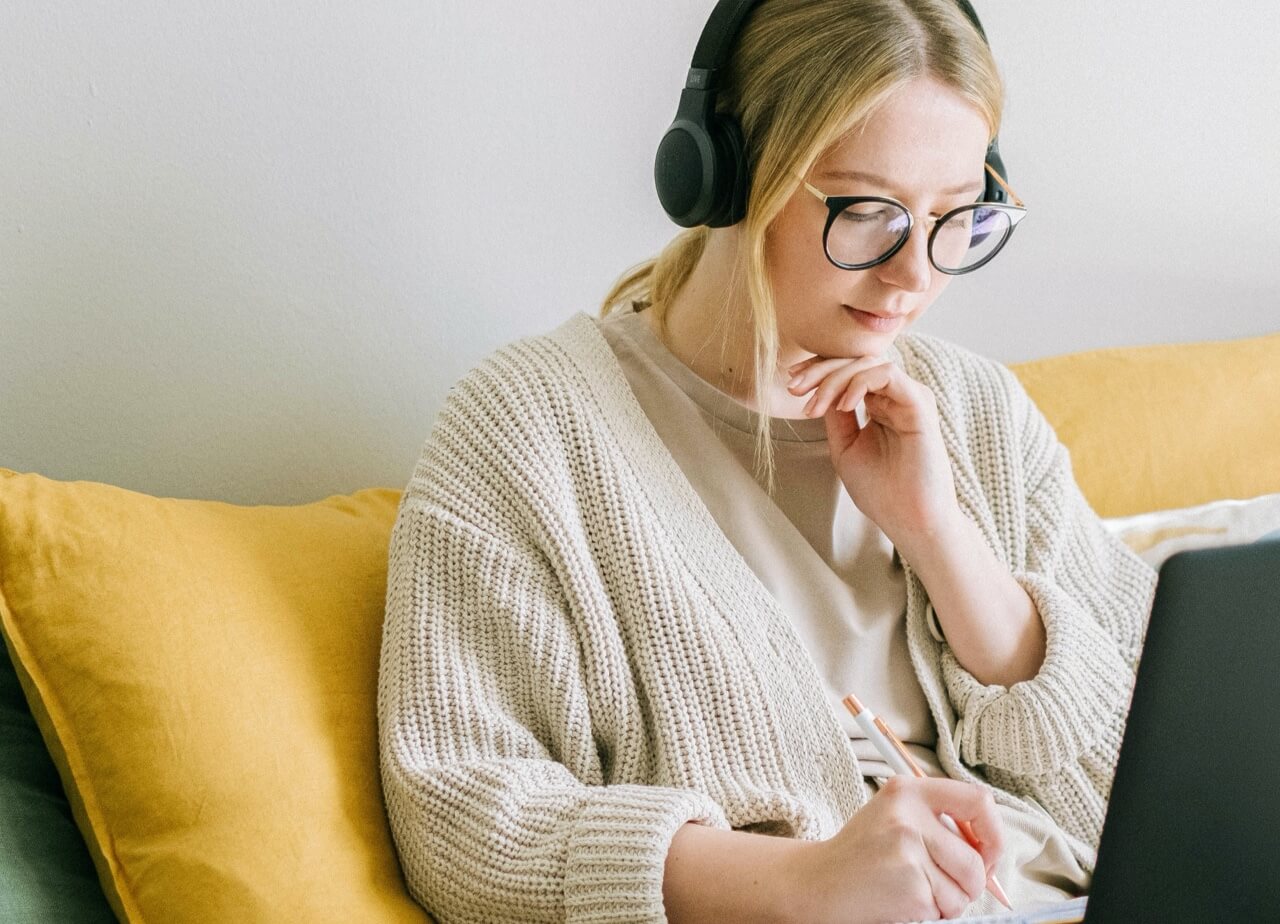 Person sitting on couch wearing wireless headphones while taking notes with a laptop in front of them.