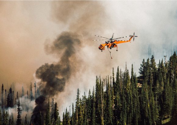 Orange firefighting helicopter flies over a smokey forest fire.