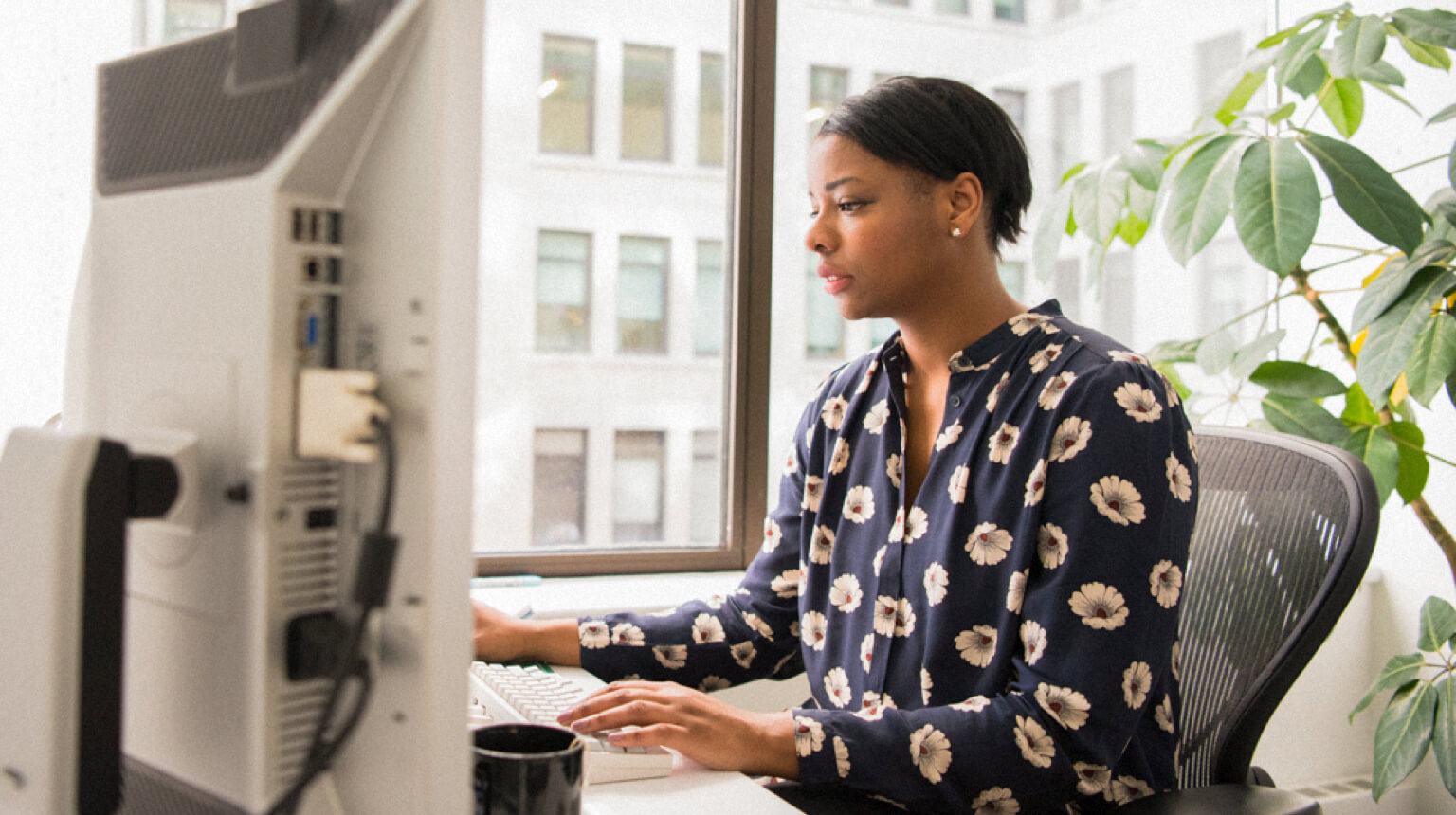 Finance professional sits at desk and works on computer next to a large window.