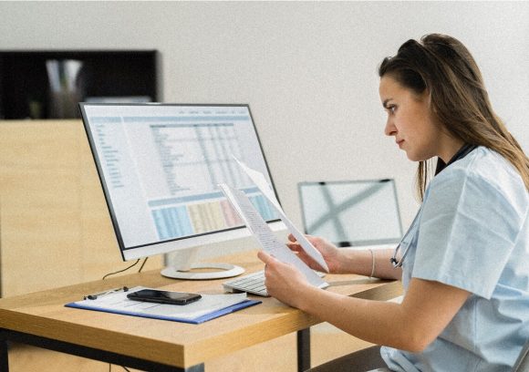 A certified medical coder sits a desk in front of a computer comparing paperwork.