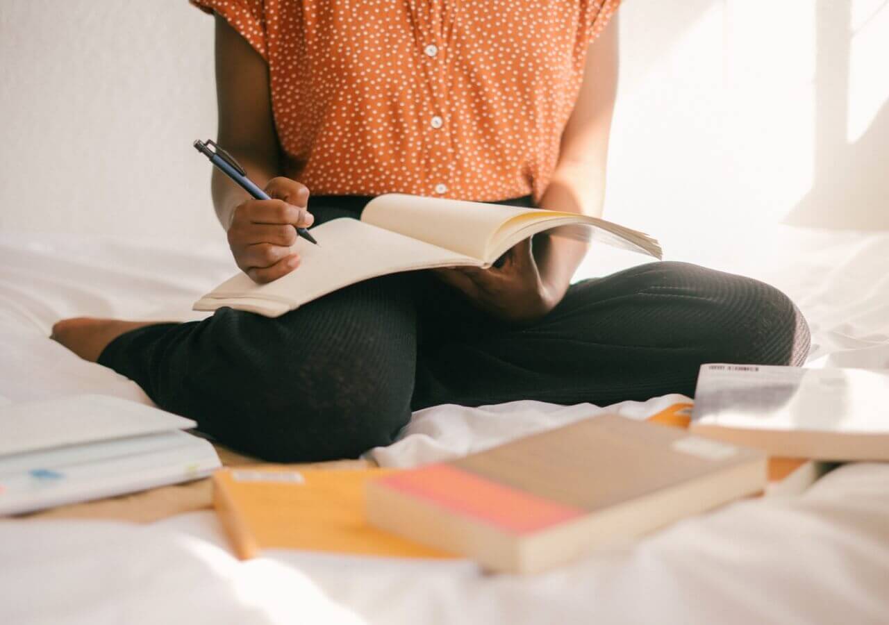 Person sitting on bed with books scattered around them while writing in a notebook.
