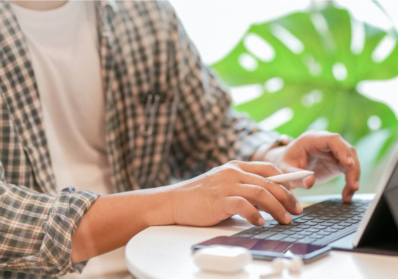 A person typing on a laptop at a table with a monstera plant in the background.