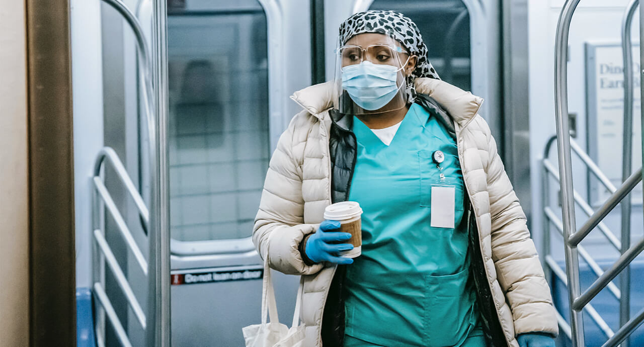 A nurse wearing a face shield and mask holding and holding a cup of coffee enters into a subway car.