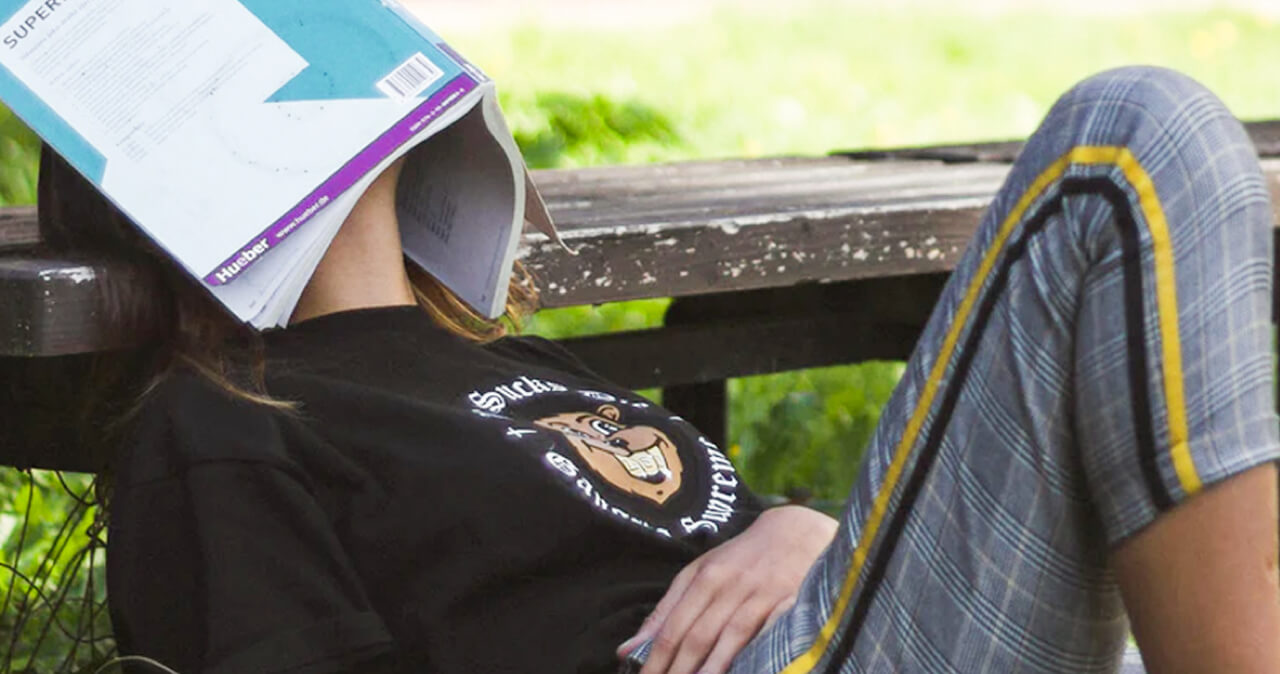 Person sitting on the ground, leaning back onto a park bench with a text book over their face.