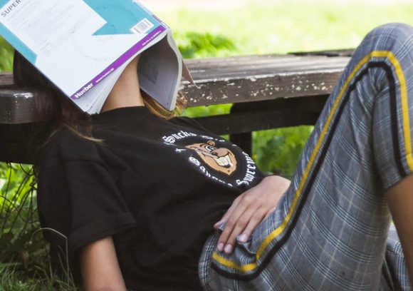 Person sitting on the ground, leaning back onto a park bench with a text book over their face.