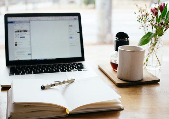 Computer on a desk with an open notebook and cup of coffee.