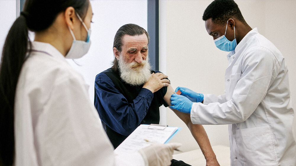 Certified clinical medical assistant putting a bandage on a patient while a doctor holding a clipboard looks on.