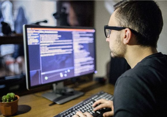 An IT professional sits at a desk with a computer while looking at lines of code.